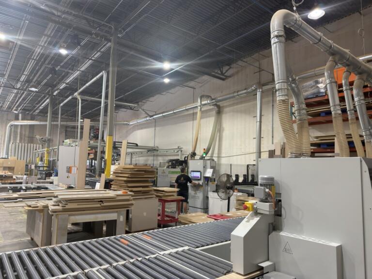 Wide view down the cabinet factory floor with overhead dust collection, roller conveyor and stacked cut parts | Sunshine Alliance, Deerfield Beach FL